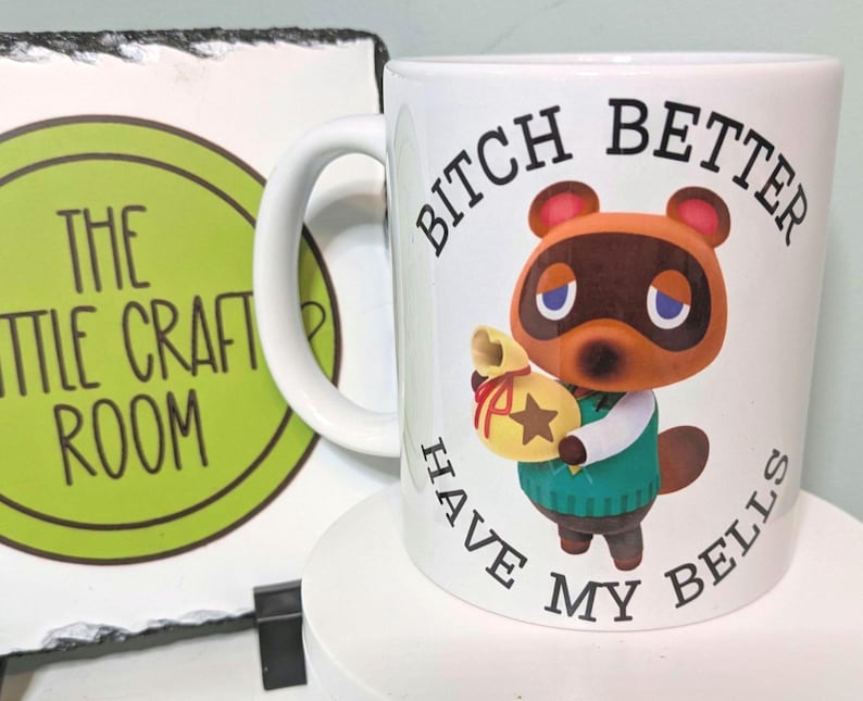 May include: A white ceramic mug featuring a graphic of the character Tom Nook from Animal Crossing holding a yellow bell bag. The text BITCH BETTER HAVE MY BELLS is printed in black capital letters around the character on the front of the mug.