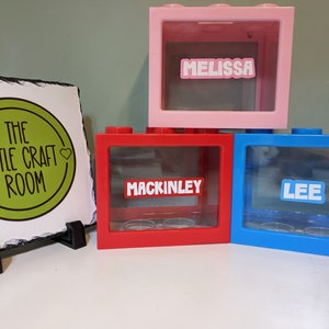 May include: Three colourful plastic money boxes with clear front panels. The money boxes are labelled "Melissa", "Mackinley", and "Lee".