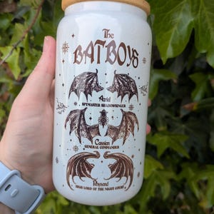 Peut inclure: Un gobelet en céramique blanc avec un couvercle et une paille en bambou. Le gobelet présente le texte "The BATBOYS" en marron, ainsi que des illustrations d'ailes de chauve-souris et les noms des personnages : Azriel, Cassian et Rhysand.