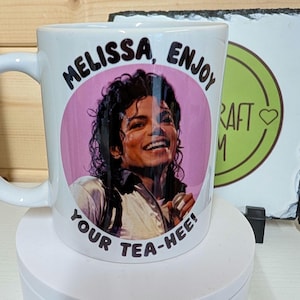 Op de afbeelding: Een witte keramische mok met een ronde roze afbeelding van een lachende Michael Jackson. De zwarte tekst rondom de afbeelding luidt "MELISSA, ENJOY YOUR TEA-HEE!". De mok heeft een gebogen handvat aan de linkerkant en staat op een witte voet.