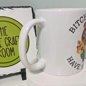 May include: A white ceramic mug featuring a cartoon raccoon character holding a yellow money bag, with the text BITCH HAVE MY MONEY printed in black capital letters. The mug is positioned next to a square slate sign with a green circle and text.