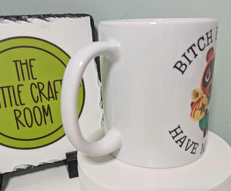 May include: A white ceramic mug featuring a cartoon raccoon character holding a yellow money bag, with the text BITCH HAVE MY MONEY printed in black capital letters. The mug is positioned next to a square slate sign with a green circle and text.