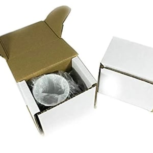 May include: Two small rectangular cardboard boxes, one open and one closed. The open box has a white exterior and brown interior flaps, containing a white ceramic mug wrapped in clear plastic. The closed box is entirely white with a lid.