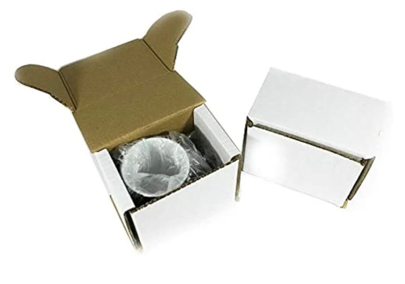 May include: Two small rectangular cardboard boxes, one open and one closed. The open box has a white exterior and brown interior flaps, containing a white ceramic mug wrapped in clear plastic. The closed box is entirely white with a lid.