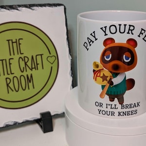 Può includere: Una tazza di ceramica bianca con un personaggio dei cartoni animati e la scritta "PAY YOUR FEES OR I'LL BREAK YOUR KNEES." Accanto, una lavagna con un cerchio verde e la scritta "THE LITTLE CRAFT ROOM."