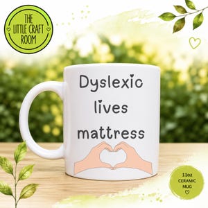 May include: A white 11oz ceramic mug featuring the black text Dyslexic lives mattress and a graphic of two hands forming a heart shape. The mug has a curved handle on the left side and is presented with a circular logo in the top left corner.