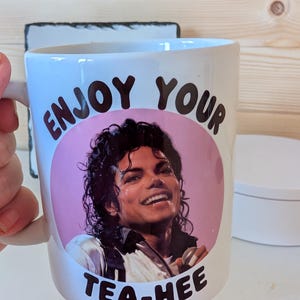 May include: A white ceramic mug featuring a circular pink graphic with a portrait of Michael Jackson. The text "ENJOY YOUR" is printed in black above the portrait, and the text "TEA-HEE" is printed in black below the portrait.