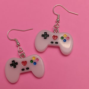 May include: A pair of light pink video game controller earrings. Each earring features a controller design with a black cross, a red heart, and colorful buttons. The earrings hang from silver-colored fish hook ear wires.