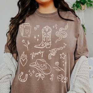 Puede incluir: Una camiseta marrón con ilustraciones blancas de estilo vaquero. El diseño incluye una bota de vaquero, un revólver, un sombrero de vaquero, una serpiente, naipes y la palabra "Howdy". La camiseta tiene un corte relajado.