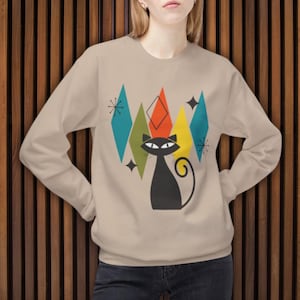 Puede incluir: Una sudadera beige con un gráfico de un gato negro. El gato está rodeado de formas geométricas coloridas en tonos de azul, verde, amarillo y naranja. El diseño tiene una estética retro moderna de mediados de siglo.