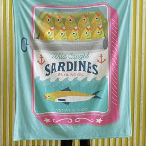 May include: A turquoise blue throw blanket with a vintage-style illustration of a can of sardines. The can has a pink and white border and says "Wild Caught Sardines in Olive Oil" with an anchor on each side. The can also has a picture of a sardine swimming in the ocean. The net weight is 3.75 oz.