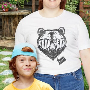 May include: A woman and a child are standing in a garden. The woman is wearing a white t-shirt with a black bear graphic and the words "Mama Bear" printed on the sunglasses. The child is wearing a yellow t-shirt and a blue cap.