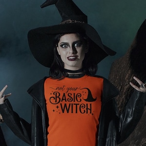 Puede incluir: Una mujer con un sombrero de bruja negro y una chaqueta de cuero negro se encuentra en un bosque oscuro. Lleva una camiseta naranja con el texto "not your basic witch" impreso.