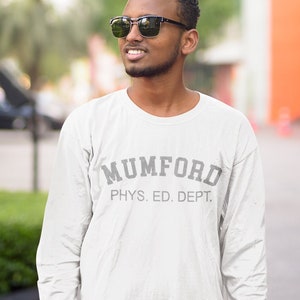 Puede incluir: Un hombre con gafas de sol y una camisa blanca de manga larga con el texto "MUMFORD PHYS. ED. DEPT." impreso en la parte delantera.