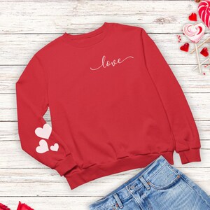 Puede incluir: Sudadera roja con la palabra "love" en escritura blanca en la parte delantera y tres corazones rosas en una manga. La sudadera se combina con una falda vaquera y zapatillas blancas.