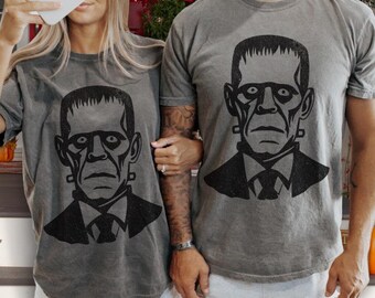 Puede incluir: Una pareja lleva camisetas grises a juego con una ilustración en blanco y negro del monstruo de Frankenstein. Las camisetas tienen un aspecto desgastado.
