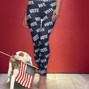 May include: A woman wearing navy blue leggings with the word "VOTE" repeated all over them. She is standing with a bulldog that is holding an American flag.