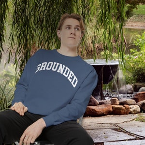 May include: A man wearing a navy blue sweatshirt with the word "GROUNDED" printed in white on the front. He is sitting on a chair in a garden setting.