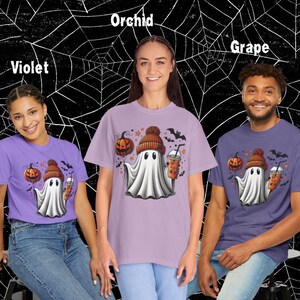 Puede incluir: Tres camisetas con un fantasma de dibujos animados que lleva un gorro y sostiene una calabaza y una bebida. Las camisetas son de color orqu&iacute;dea, violeta y uva.