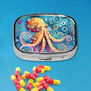 May include: A silver pill box with a colorful octopus design. The octopus is orange, yellow, and purple against a blue background. Red and yellow capsules are scattered around the box.