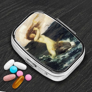 May include: A silver pill box with a hinged lid, featuring a classical painting of a nude woman in the sea. Assorted pills in various colors are scattered beside the box. The box is rectangular with rounded corners.
