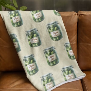 May include: A soft, beige fleece throw blanket with a repeating pattern of green pickle jars labelled "Pickle Salad".
