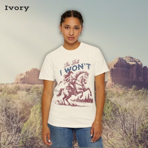 May include: A white t-shirt with a vintage-style graphic of a woman riding a horse. The graphic says "The Hell I Won't".