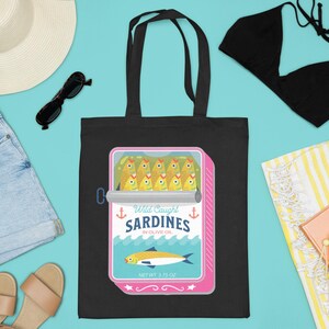 May include: Black tote bag with a graphic of a pink can of sardines. The can has a white label with the text "Wild Caught Sardines in Olive Oil" and a picture of a sardine. The bag is on a blue background with other beach-themed items.