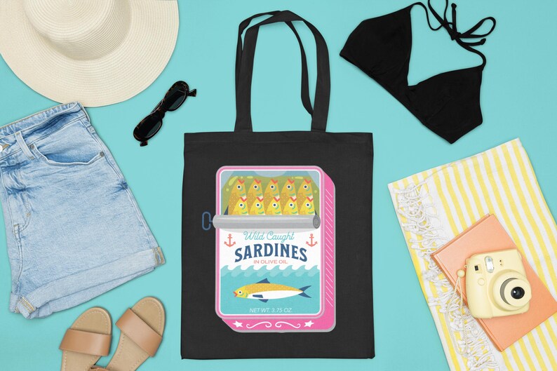 May include: Black tote bag with a graphic of a pink can of sardines. The can has a white label with the text "Wild Caught Sardines in Olive Oil" and a picture of a sardine. The bag is on a blue background with other beach-themed items.