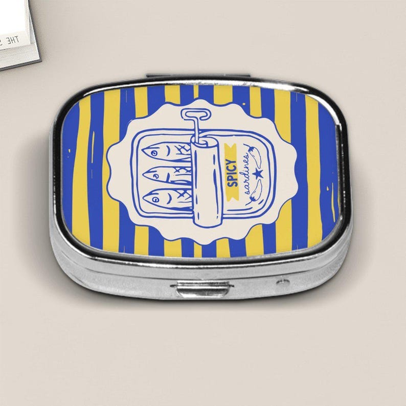 Peut inclure: Une bo&icirc;te &agrave; pilules argent&eacute;e avec un motif &agrave; rayures bleues et jaunes. Le couvercle pr&eacute;sente une &eacute;tiquette blanche avec une illustration bleue de sardines en bo&icirc;te, avec le texte "SPICY sardines".