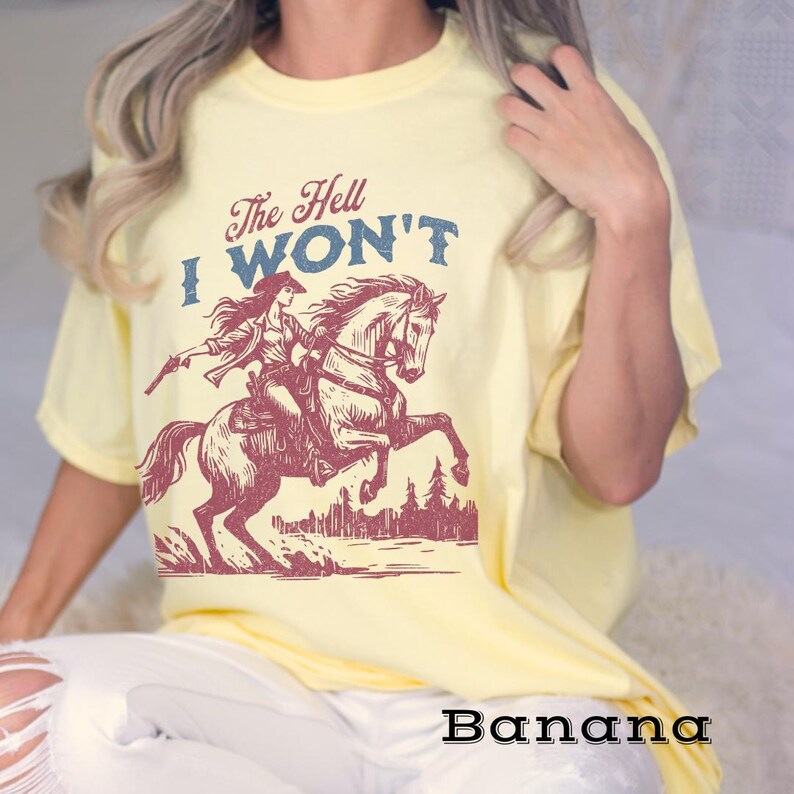 May include: A yellow t-shirt with a vintage-style graphic of a woman riding a horse. The text "The Hell I Won't" is printed on the shirt in a red and blue font.