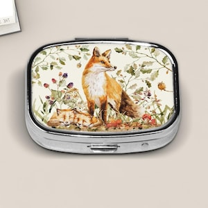 May include: A silver pill box with a watercolor illustration of a fox family in a forest setting. The design features a standing fox and two sleeping foxes, surrounded by foliage, mushrooms, and berries. The box has a rectangular shape with rounded edges.