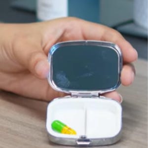 Puede incluir: Pastillero plateado abierto con espejo en la tapa. El interior est&aacute; dividido en dos compartimentos, uno de los cuales contiene una c&aacute;psula verde y amarilla. El estuche est&aacute; abierto por una mano.
