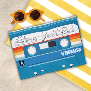 May include: A blue cassette tape-shaped pouch with the words "Yacht Rock" and "Vintage" printed on it. The pouch has a retro design with a boat illustration and a striped rainbow. Orange sunglasses rest on a yellow and white striped towel.