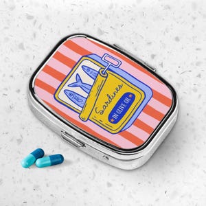 Peut inclure: Une bo&icirc;te &agrave; pilules argent&eacute;e avec un motif de sardines. Le couvercle pr&eacute;sente un fond ray&eacute; rose et orange avec une illustration de bo&icirc;te de sardines jaune. La bo&icirc;te indique "Sardines in Olive Oil". Deux pilules bleues et turquoise sont &agrave; c&ocirc;t&eacute; de la bo&icirc;te.