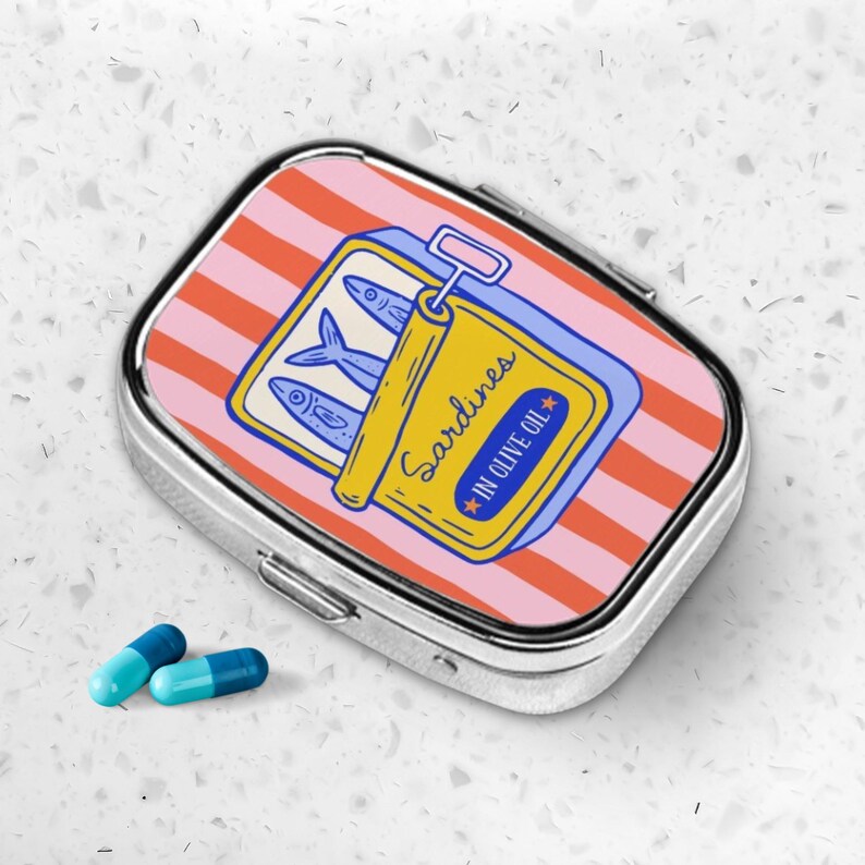 Peut inclure: Une bo&icirc;te &agrave; pilules argent&eacute;e avec un motif de sardines. Le couvercle pr&eacute;sente un fond ray&eacute; rose et orange avec une illustration de bo&icirc;te de sardines jaune. La bo&icirc;te indique "Sardines in Olive Oil". Deux pilules bleues et turquoise sont &agrave; c&ocirc;t&eacute; de la bo&icirc;te.