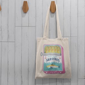 May include: A white canvas tote bag with a colorful illustration of a can of sardines. The can has a pink label with the text "Wild Caught Sardines" and a blue and white illustration of a fish.