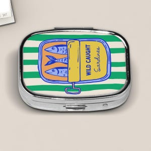 May include: A silver pill box with a sardine can design. The sardine can is yellow with the words "WILD CAUGHT Sardines" and features three blue fish. The background has green and white stripes.