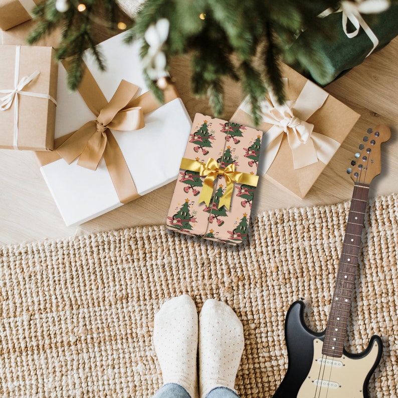 May include: A pink gift box with a gold ribbon and a pattern of green Christmas trees. The box is sitting on a brown rug with a guitar and a pair of white socks in the foreground.