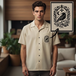 May include: A short-sleeved, beige button-down shirt with a black raven graphic on the pocket. The graphic features a raven perched on a branch with roses, a crescent moon, and a cross. The shirt has a classic collar and black buttons.