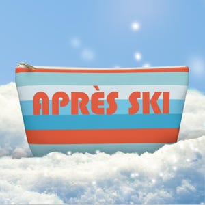 May include: A rectangular cosmetic bag with a gold zip and the words "APRÈS SKI" in orange block letters. The bag has horizontal stripes in light blue, white, and orange. The background is a sky with clouds.