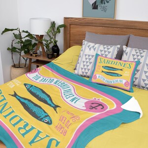 Puede incluir: Un juego de manta y almohada amarillas con un dise&ntilde;o de sardinas. La manta presenta dos peces azules y el texto "Sardinas en aceite de oliva virgen extra". La almohada tiene el mismo dise&ntilde;o. El juego est&aacute; sobre una cama amarilla.
