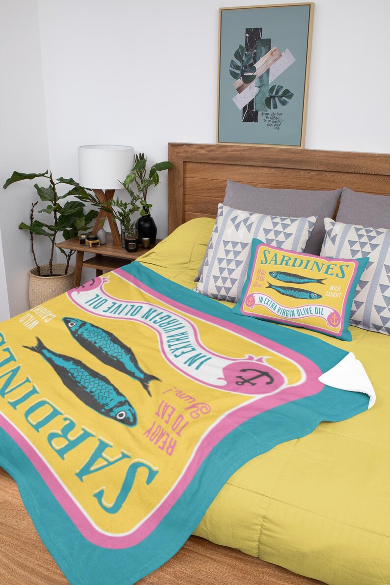Puede incluir: Un juego de manta y almohada amarillas con un dise&ntilde;o de sardinas. La manta presenta dos peces azules y el texto "Sardinas en aceite de oliva virgen extra". La almohada tiene el mismo dise&ntilde;o. El juego est&aacute; sobre una cama amarilla.