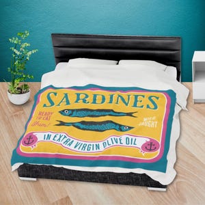 Puede incluir: Una manta con un dise&ntilde;o de etiqueta de sardinas de estilo vintage. La etiqueta es amarilla con detalles en azul y rosa, con dos ilustraciones de peces y el texto "Sardinas en aceite de oliva virgen extra."