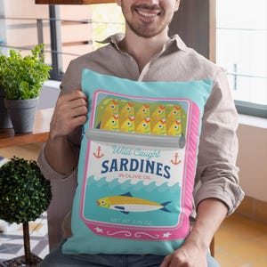 May include: A turquoise blue cushion with a pink border features a vintage-style illustration of a can of sardines. The can has a white label with the text "Wild Caught Sardines in Olive Oil" and "Net Wt. 3.75 oz."