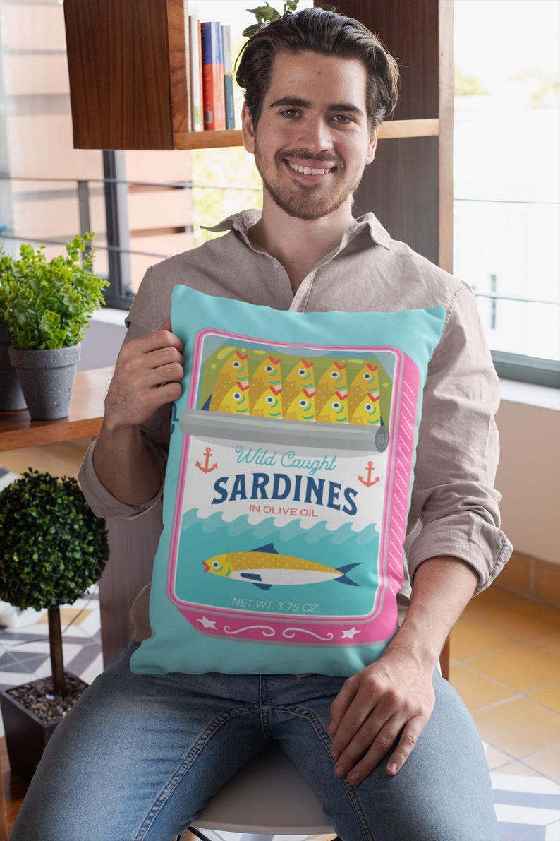 May include: A turquoise blue cushion with a pink border features a vintage-style illustration of a can of sardines. The can has a white label with the text "Wild Caught Sardines in Olive Oil" and "Net Wt. 3.75 oz."
