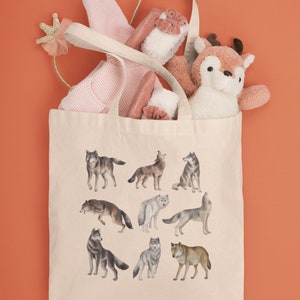 May include: A beige canvas tote bag featuring a design of various wolves in different poses. The bag is filled with a pink sweater, a stuffed deer, and a pink headband with a gold crown.