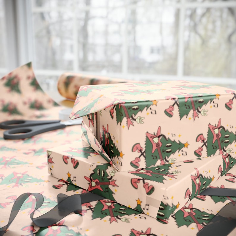 May include: Two pink gift boxes wrapped in Christmas wrapping paper with a pattern of green Christmas trees playing electric guitars. The boxes are stacked on top of each other and are tied with a black ribbon.