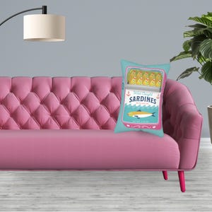 May include: A pink sofa with a blue and white cushion featuring a cartoon illustration of a can of sardines. The can says "Wild Caught Sardines in Olive Oil" and has a net weight of 2.25 oz.