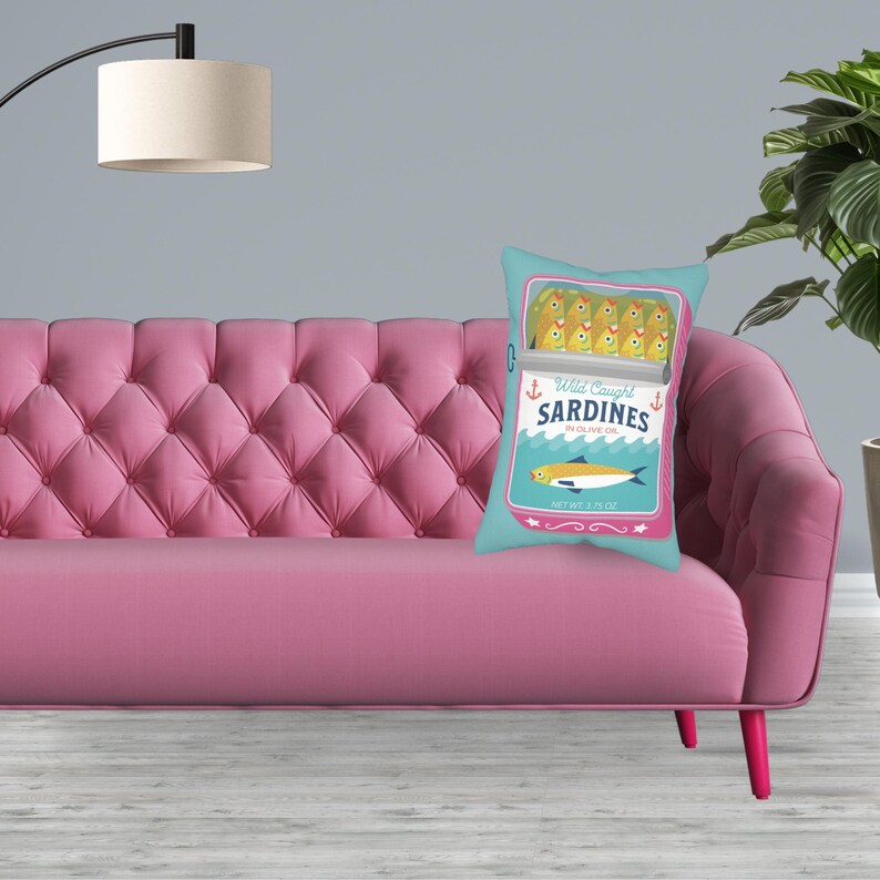 May include: A pink sofa with a blue and white cushion featuring a cartoon illustration of a can of sardines. The can says "Wild Caught Sardines in Olive Oil" and has a net weight of 2.25 oz.